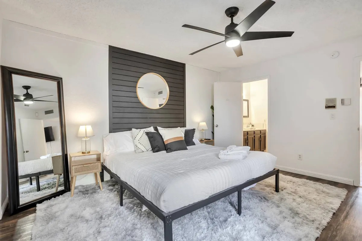 This room features a serene bedroom with a cozy bed, decorative mirror, and a nightstand with a lamp. The presence of natural light and soft textures creates an inviting atmosphere ideal for privacy and relaxation.