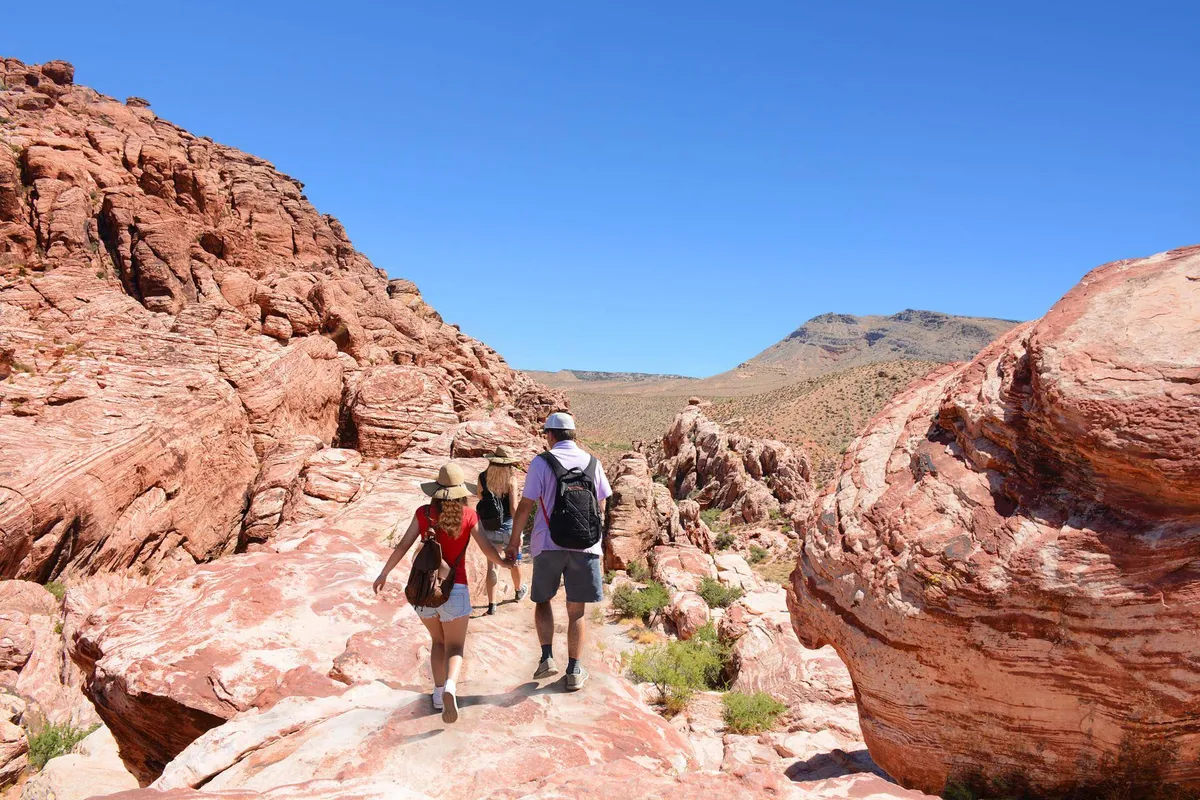 This image captures a striking exterior scene of red rock formations under a bright blue sky, with hikers exploring the landscape. The composition emphasizes adventure, natural beauty, and the spirit of discovery.