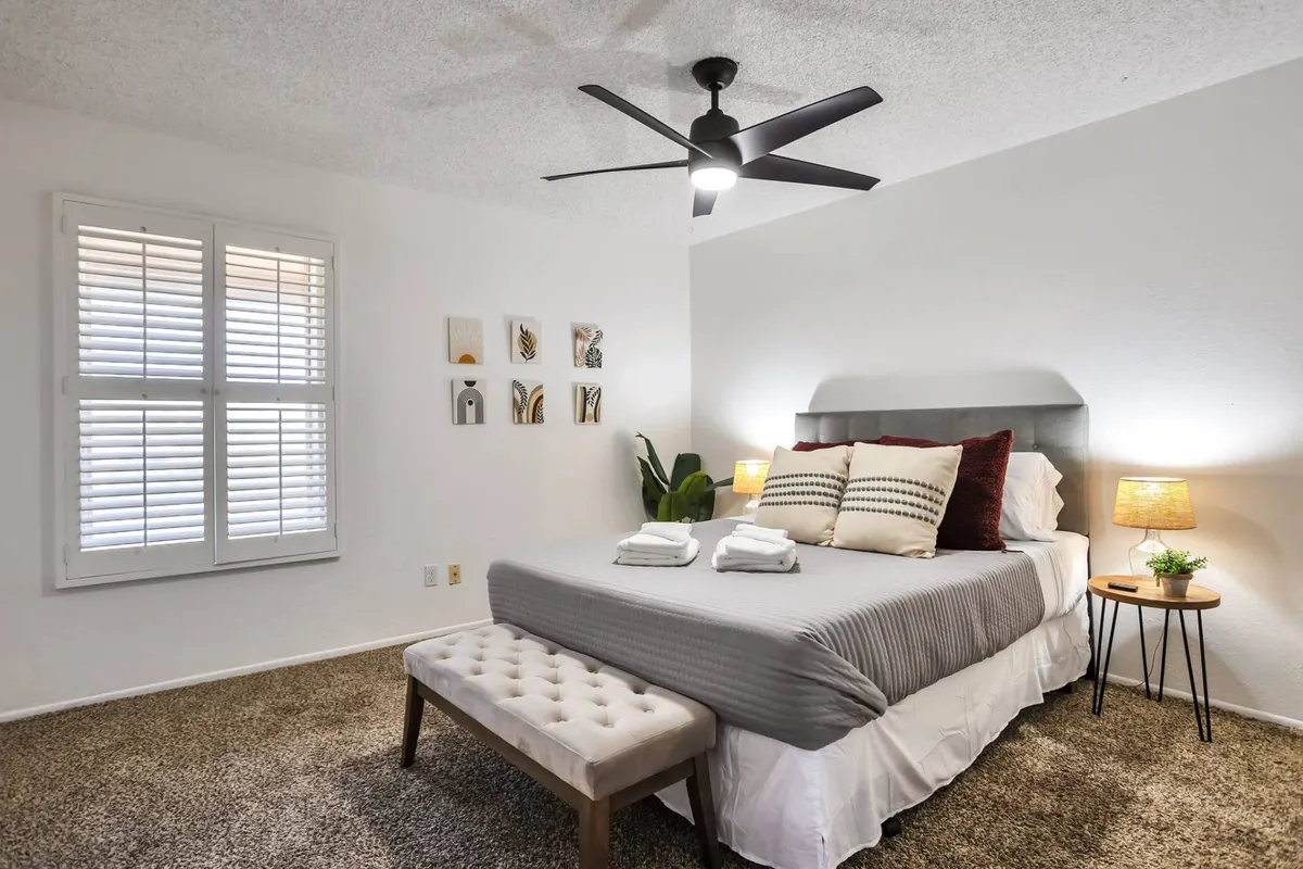 A tranquil master bedroom featuring a large bed with decorative pillows, matching nightstands with lamps on either side, framed wall art above the headboard, window shutters letting in gentle light, and a leafy plant in the corner. The setting feels cozy and restful, ideal for relaxation or a quiet read.