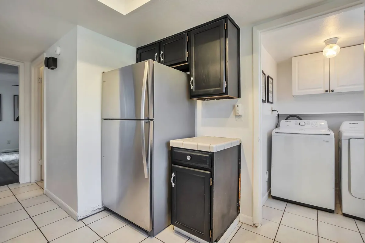 The image highlights a practical laundry area adjacent to the kitchen, featuring a modern washer and dryer set, refrigerator, ample counter space, and a tidy environment. The setup suggests easy multitasking between daily chores and meal preparation.