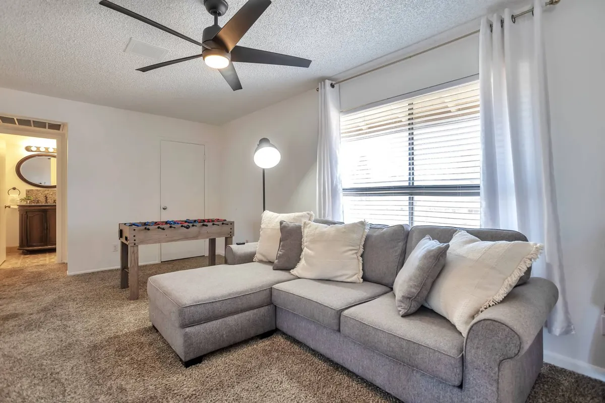 This image presents a bright upstairs loft area featuring a large sectional sofa, a foosball table, a floor lamp, and a large window that brings in ample natural light. The playful arrangement creates a comfortable setting for social gatherings, games, and relaxation.