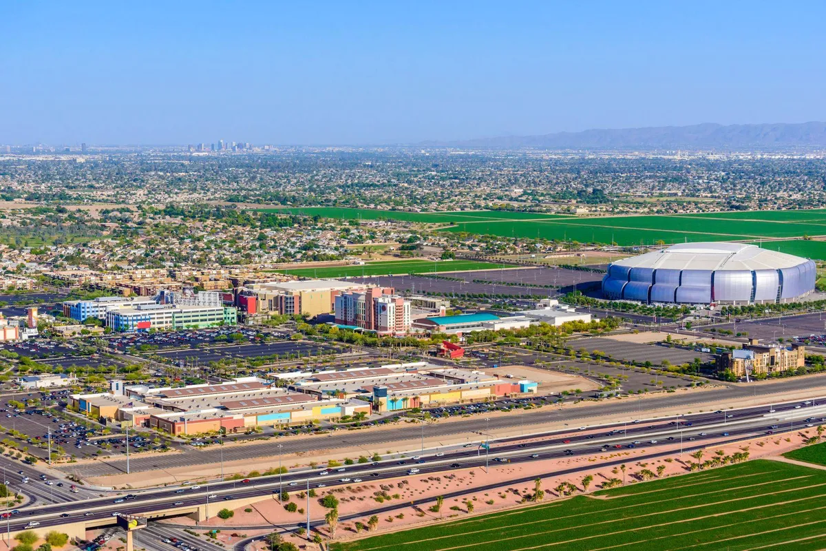 The image captures a broad view of the neighborhood with a large stadium, commercial buildings, open green fields, and a highway, illustrating the vibrant surroundings and the variety of activities available nearby.