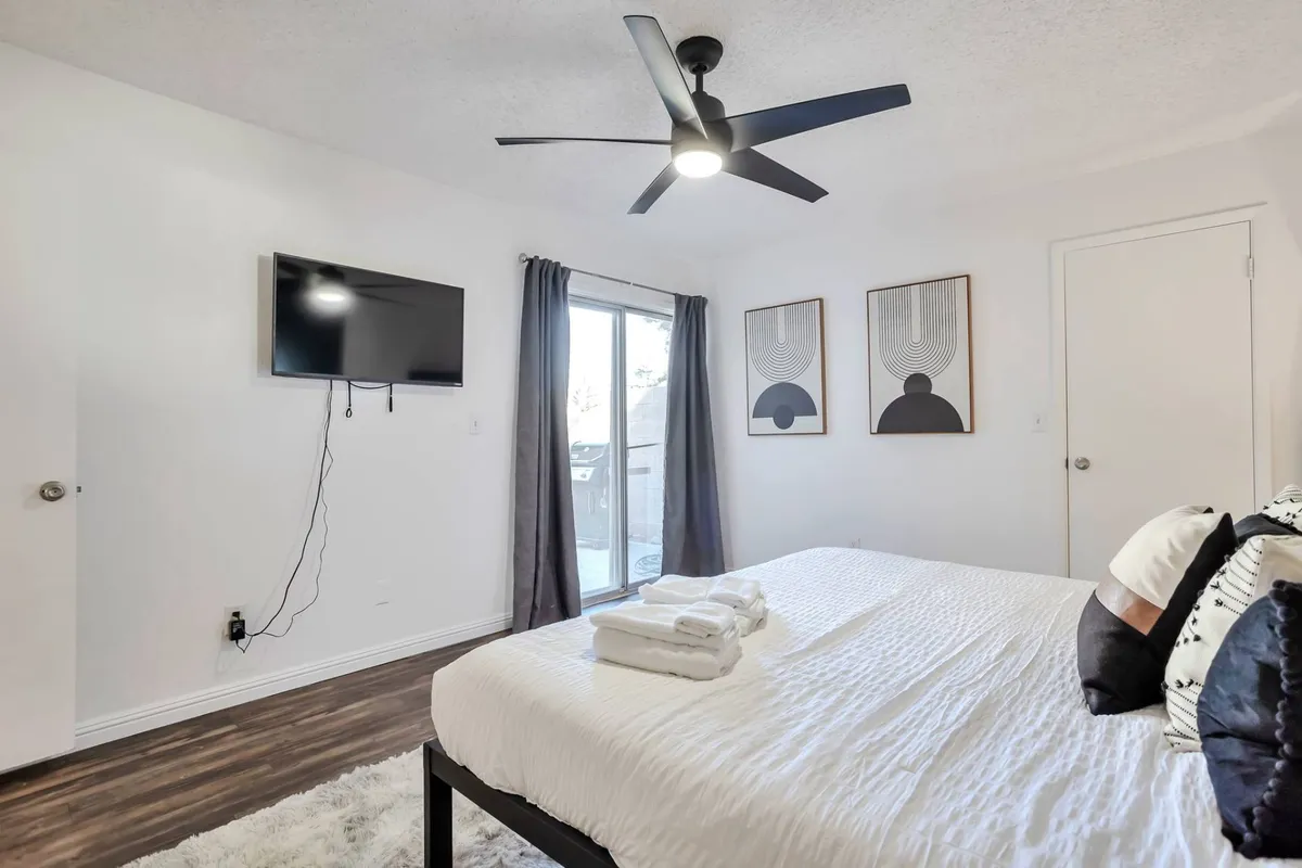 A tranquil bedroom with crisp white linens, wall-mounted TV, tasteful artwork, and access to a balcony. The open balcony door suggests a seamless connection between indoor comfort and outdoor views, enhancing the sense of retreat.