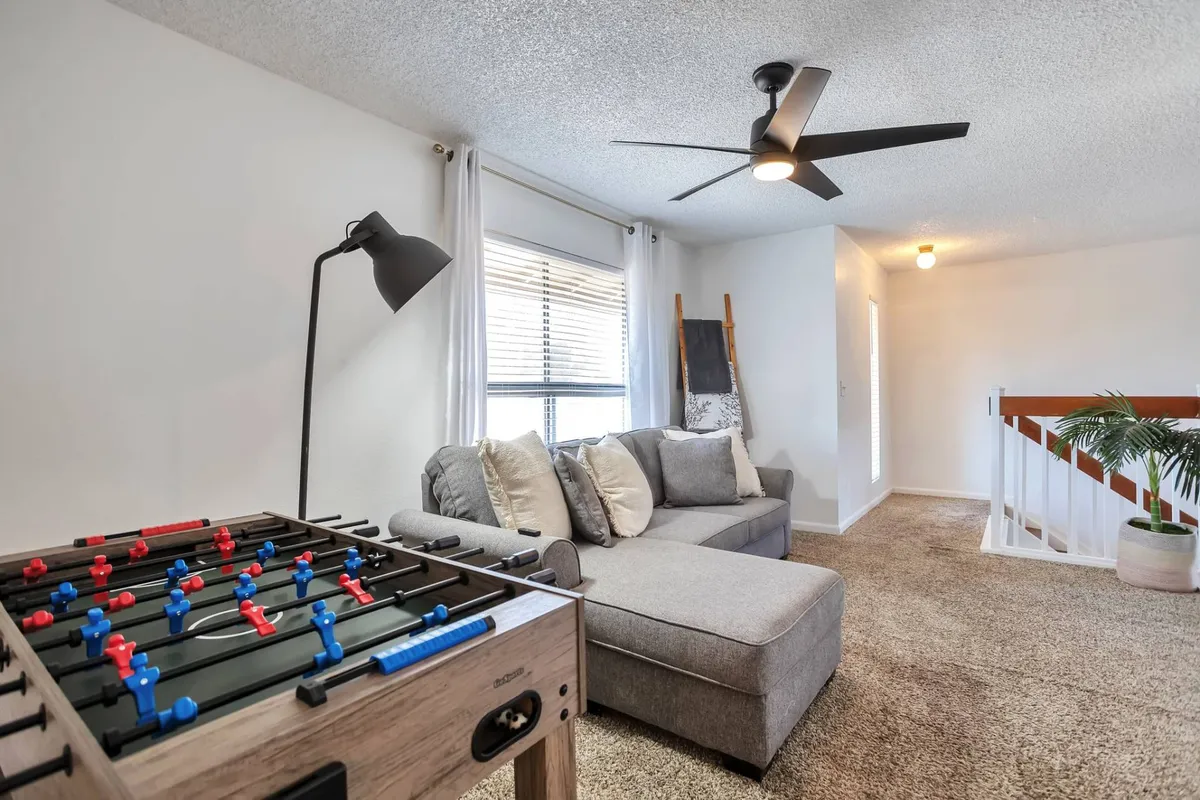 A bright, playful living area featuring a foosball table, large comfortable sofa, ceiling fan, and ample natural light streaming through big windows. The space is set up for socializing, with plenty of room for friends to gather, play games, or relax together after a sporting event.