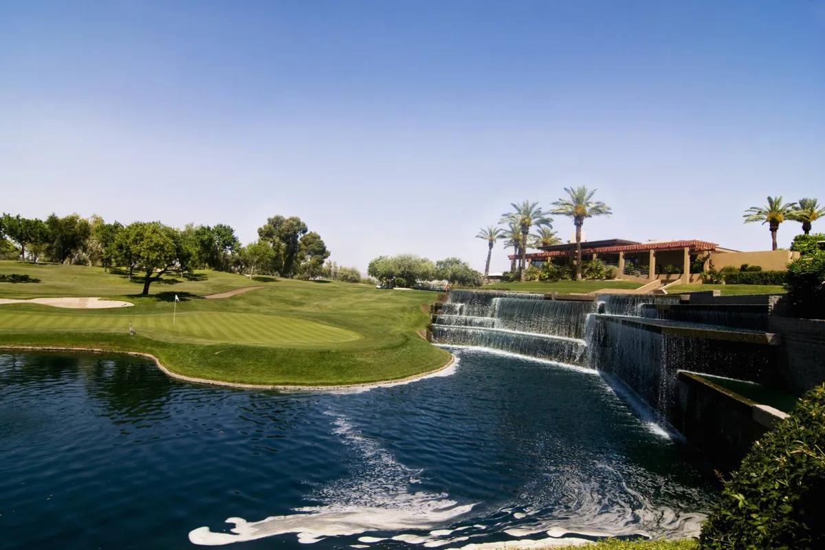 A sweeping view of the Bellair Golf Course with manicured grass, a tranquil waterfall, palm trees, and a clear blue sky. The image exudes relaxation and the vibrant outdoor lifestyle available just steps from the property.